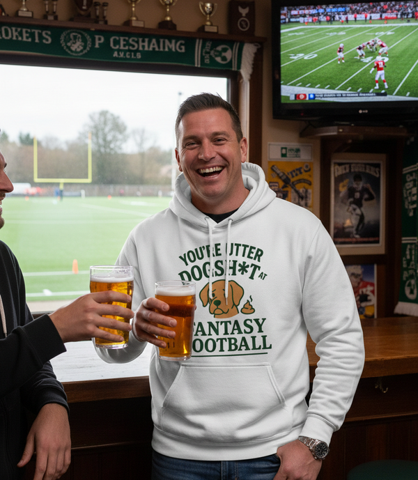 Man wearing a hoodie with humorous fantasy football text, holding beer glasses in a pub setting with sports memorabilia. The man is wearing a Cows Corner hoodie with the funny fantasy football slogan on the front that reads 'You're Utter Dogsh*t at Fantasy Football'.  Cows Corner gifts are perfect for sport-mad fans, these gifts work brilliantly for birthdays, new baby celebrations, Father’s Day, Mother’s Day, Christmas, anniversaries, thank you gifts, end-of-season team awards, graduations, retirements