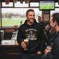 Man in a bar wearing a humorous hoodie, holding a beer, with a TV showing a sports game in the background.  The man is wearing a Cows Corner hoodie with the funny fantasy football slogan on the front that reads 'Your Fantasy Team Has One Setting: Utter Sh*tness'. Cows Corner gifts are perfect for sport-mad fans, these gifts work brilliantly for birthdays, new baby celebrations, Father’s Day, Mother’s Day, Christmas, anniversaries, thank you gifts, end-of-season team awards, graduations, retirements