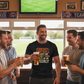 Group of men in a pub watching a football match on TV, one wearing a humorous 'Fantasy Football' t-shirt. The man is wearing a Cows Corner t-shirt with the funny fantasy football slogan on the front that reads 'Fantasy Football Is Just Me Getting F*cked Over Weekly'. Cows Corner gifts are perfect for sport-mad fans, these gifts work brilliantly for birthdays, new baby celebrations, Father’s Day, Mother’s Day, Christmas, anniversaries, thank you gifts, end-of-season team awards, graduations, retirements