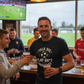 Man in a bar holding a beer with others, watching sports on TV. Man wearing a Cows Corner t-shirt with the funny fantasy football slogan on the front that reads 'Talk Big, Play Sh*t, Finish Last'.  Cows Corner gifts are perfect for sport-mad fans, these gifts work brilliantly for birthdays, new baby celebrations, Father’s Day, Mother’s Day, Christmas, anniversaries, thank you gifts, end-of-season team awards, graduations, retirements, and just-because moments when you want to raise a smile. 