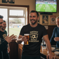 Man holding a beer in a pub with a television showing a sports event in the background, the man is wearing a Cows Corner t-shirt with the funny fantasy football slogan on the front that reads 'Fantasy Football = Weekly Humiliation'. Cows Corner gifts are perfect for sport-mad fans, these gifts work brilliantly for birthdays, new baby celebrations, Father’s Day, Mother’s Day, Christmas, anniversaries, thank you gifts, end-of-season team awards, graduations, retirements, and just-because moments 