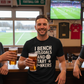 Man holding a beer in a pub with a TV showing a football match in the background. The man is wearing a Cows Corner t-shirt with the funny fantasy football slogan on the front that reads 'I Bench Goals and Start W*nkers'. Cows Corner gifts are perfect for sport-mad fans, these gifts work brilliantly for birthdays, new baby celebrations, Father’s Day, Mother’s Day, Christmas, anniversaries, thank you gifts, end-of-season team awards, graduations, retirements, and just-because moments 
