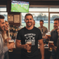 Group of people in a bar watching sports on TV, one person wearing a humorous t-shirt.  Man wearing a Cows Corner hoodie with the funny fantasy football slogan on the front that reads 'My Fantasy Team Is F*cking Useless'. Cows Corner gifts are perfect for sport-mad fans, these gifts work brilliantly for birthdays, new baby celebrations, Father’s Day, Mother’s Day, Christmas, anniversaries, thank you gifts, end-of-season team awards, graduations, retirements, and just-because moments 