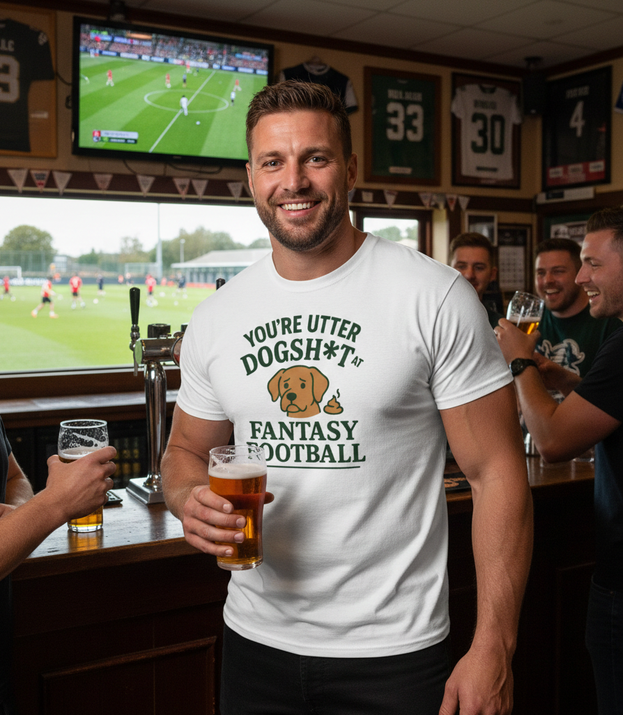 Man in a pub wearing a humorous t-shirt holding a beer, with sports jerseys on the wall. The man is wearing a Cows Corner T-shirt with the funny fantasy football slogan on the front that reads 'You're Utter Dogsh*t at Fantasy Football'. Cows Corner gifts are perfect for sport-mad fans, these gifts work brilliantly for birthdays, new baby celebrations, Father’s Day, Mother’s Day, Christmas, anniversaries, thank you gifts, end-of-season team awards, graduations, retirements, and just-because moments 