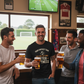 Four men in a pub watching a soccer match on television, holding beer glasses.  The man is wearing a Cows Corner T-shirt with the funny fantasy football slogan on the front that reads 'Your Fantasy Team Has One Setting: Utter Sh*tness'. Cows Corner gifts are perfect for sport-mad fans, these gifts work brilliantly for birthdays, new baby celebrations, Father’s Day, Mother’s Day, Christmas, anniversaries, thank you gifts, end-of-season team awards, graduations, retirements, and just-because moments 