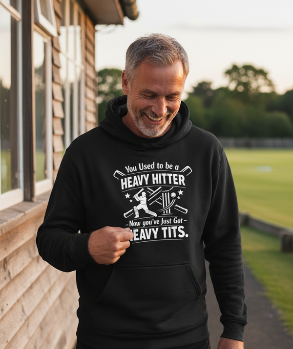 Man wearing a black hoodie with humorous text outdoors. The man is wearing a Cows Corner hoodie with a funny cricket slogan 'You Used to be a Heavy Hitter. Now you’ve Just Got Heavy Tits'. Cows Corner gifts are perfect for sport-mad fans, these gifts work brilliantly for birthdays, new baby celebrations, Father’s Day, Mother’s Day, Christmas, anniversaries, thank you gifts, end-of-season team awards, graduations, retirements, and just-because moments when you want to raise a smile. 
