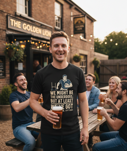 Man holding a beer in front of 'The Golden Lion' pub with friends around him.  The man is wearing a Cows Corner t-shirt with the funny rugby slogan 'We Might Be the Underdogs, but at Least We’ve Got Better Teeth – Scott Gibbs'. Cows Corner gifts are perfect for sport-mad fans, these gifts work brilliantly for birthdays, new baby celebrations, Father’s Day, Mother’s Day, Christmas, anniversaries, thank you gifts, end-of-season team awards, graduations, retirements, and just-because moments 
