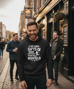 Man wearing a black hoodie with humorous text on a street scene. The man is wearing a Cows Corner t-shirt with the funny football slogan 'I’ve Had Sh*ts More Impressive Than My Midfield'. Cows Corner gifts are perfect for sport-mad fans, these gifts work brilliantly for birthdays, new baby celebrations, Father’s Day, Mother’s Day, Christmas, anniversaries, thank you gifts, end-of-season team awards, graduations, retirements, and just-because moments when you want to raise a smile. 
