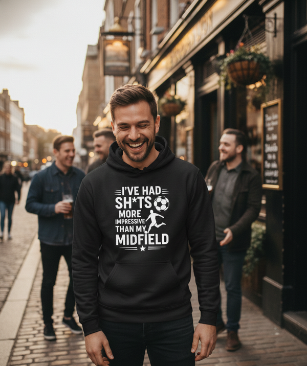 Man wearing a black hoodie with humorous text on a street scene. The man is wearing a Cows Corner t-shirt with the funny football slogan 'I’ve Had Sh*ts More Impressive Than My Midfield'. Cows Corner gifts are perfect for sport-mad fans, these gifts work brilliantly for birthdays, new baby celebrations, Father’s Day, Mother’s Day, Christmas, anniversaries, thank you gifts, end-of-season team awards, graduations, retirements, and just-because moments when you want to raise a smile. 
