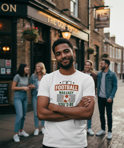 Man wearing a white t-shirt with a football design standing on a street with people in the background. The man is wearing a Cows Corner t-shirt with the funny football slogan 'If Football Was Easy You’d Be Good at It'.  Cows Corner gifts are perfect for sport-mad fans, these gifts work brilliantly for birthdays, new baby celebrations, Father’s Day, Mother’s Day, Christmas, anniversaries, thank you gifts, end-of-season team awards, graduations, retirements, and just-because moments 