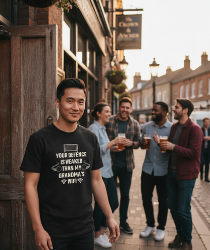 Man wearing a black t-shirt with text standing on a street with friends in the background. The man is wearing a Cows Corner t-shirt with the funny football slogan 'Your Defence Is Weaker Than My Grandma’s Wifi'. Cows Corner gifts are perfect for sport-mad fans, these gifts work brilliantly for birthdays, new baby celebrations, Father’s Day, Mother’s Day, Christmas, anniversaries, thank you gifts, end-of-season team awards, graduations, retirements, and just-because moments when you want to raise a smile. 
