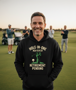 Man wearing a black hoodie with a humorous retirement message on a golf course.  The man is wearing a Cows Corner hoodie with the funny golf slogan 'Hole-In-One Achieved… Retirement Pending'. Cows Corner gifts are perfect for sport-mad fans, these gifts work brilliantly for birthdays, new baby celebrations, Father’s Day, Mother’s Day, Christmas, anniversaries, thank you gifts, end-of-season team awards, graduations, retirements, and just-because moments when you want to raise a smile. 

