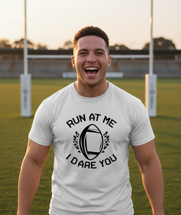 Man wearing a white t-shirt with a rugby ball design and text on a sports field. The man is wearing a Cows Corner t-shirt with the funny rugby slogan 'Run at Me I Dare You'. Cows Corner gifts are perfect for sport-mad fans, these gifts work brilliantly for birthdays, new baby celebrations, Father’s Day, Mother’s Day, Christmas, anniversaries, thank you gifts, end-of-season team awards, graduations, retirements, and just-because moments when you want to raise a smile. 
