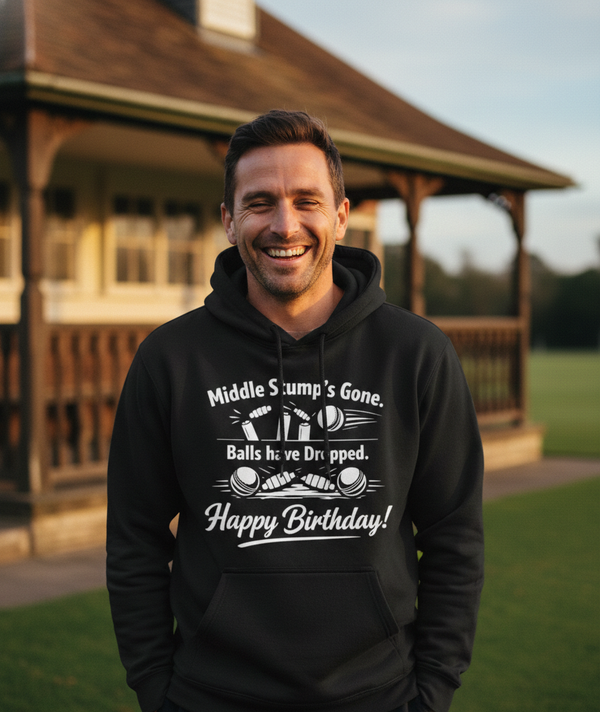 Man wearing a black hoodie with humorous text in front of a wooden building. The man is wearing a Cows corner hoodie with the funny cricket slogan 'Middle Stump’s Gone. Balls have Dropped. Happy Birthday'. Cows Corner gifts are perfect for sport-mad fans, these gifts work brilliantly for birthdays, new baby celebrations, Father’s Day, Mother’s Day, Christmas, anniversaries, thank you gifts, end-of-season team awards, graduations, retirements, and just-because moments when you want to raise a smile. 
