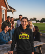 Person wearing a black hoodie with text, standing outdoors with friends in the background. The lady is wearing a Cows Corner hoodie with a funny cricket slogan 'Mind The Gap Between Your Bat And Pads'. Cows Corner gifts are perfect for sport-mad fans, these gifts work brilliantly for birthdays, new baby celebrations, Father’s Day, Mother’s Day, Christmas, anniversaries, thank you gifts, end-of-season team awards, graduations, retirements, and just-because moments when you want to raise a smile. 
