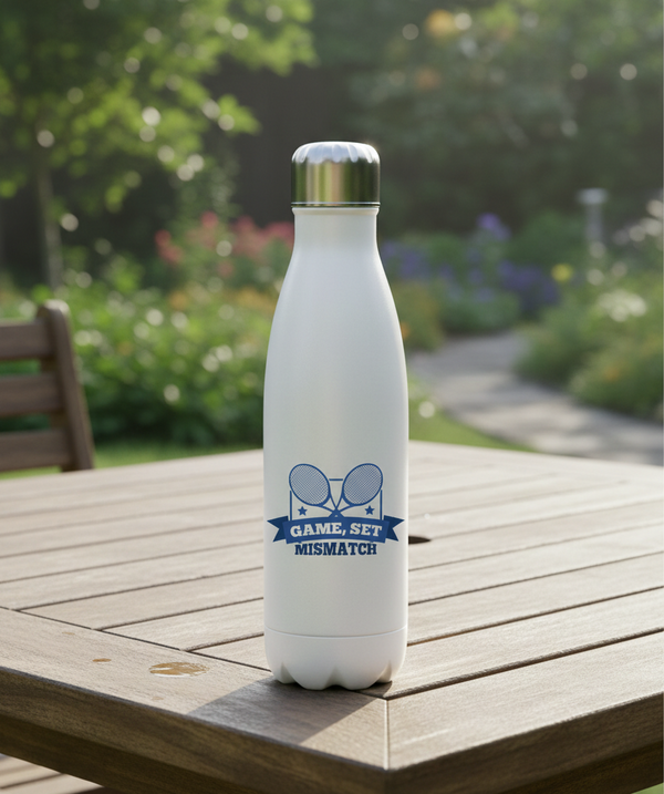 White water bottle with a logo on a wooden table outdoors. This is a Cows Corner water bottle with a funny tennis slogan 'Game, Set, Mismatch'. Cows Corner gifts are perfect for sport-mad fans, these gifts work brilliantly for birthdays, new baby celebrations, Father’s Day, Mother’s Day, Christmas, anniversaries, thank you gifts, end-of-season team awards, graduations, retirements, and just-because moments when you want to raise a smile. 
