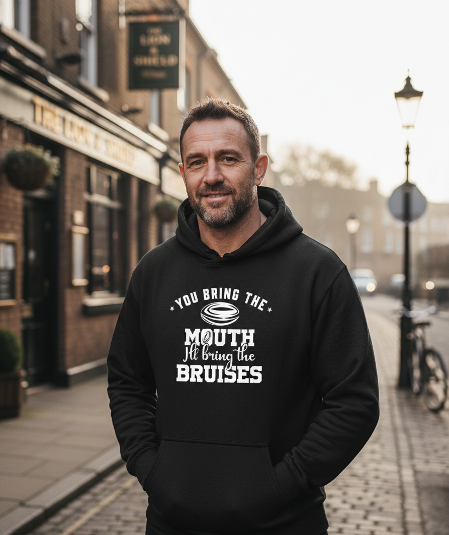 Man wearing a black hoodie with text on a street. The man is wearing a Cows Corner hoodie with a funny rugby slogan 'You Bring the Mouth I’ll Bring the Bruises'. Cows Corner gifts are perfect for sport-mad fans, these gifts work brilliantly for birthdays, new baby celebrations, Father’s Day, Mother’s Day, Christmas, anniversaries, thank you gifts, end-of-season team awards, graduations, retirements, and just-because moments when you want to raise a smile. 
