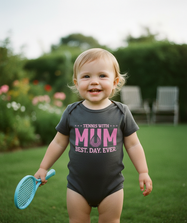 Child wearing a onesie with 'Tennis with Mum Best Day Ever' text, holding a blue racket outdoors. Cows Corner gifts are perfect for sport-mad fans, these gifts work brilliantly for birthdays, new baby celebrations, Father’s Day, Mother’s Day, Christmas, anniversaries, thank you gifts, end-of-season team awards, graduations, retirements, and just-because moments when you want to raise a smile. 
