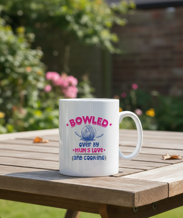 White mug with colorful text on a wooden table outdoors. This is a Cows Corner mug with a funny cricket slogan 'Bowled Over by My Mum’s Love & Cooking'. Cows Corner gifts are perfect for sport-mad fans, these gifts work brilliantly for birthdays, new baby celebrations, Father’s Day, Mother’s Day, Christmas, anniversaries, thank you gifts, end-of-season team awards, graduations, retirements, and just-because moments when you want to raise a smile. 
