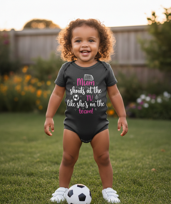 Child wearing a black onesie with text outdoors on grass. The baby is wearing a Cows Corner baby grow with the funny football slogan 'Mum Shouts at the TV Like She’s on the Team!'. Cows Corner gifts are perfect for sport-mad fans, these gifts work brilliantly for birthdays, new baby celebrations, Father’s Day, Mother’s Day, Christmas, anniversaries, thank you gifts, end-of-season team awards, graduations, retirements, and just-because moments when you want to raise a smile. 

