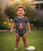 Child wearing a onesie with text and a cartoon character, standing on grass with flowers in the background. The baby is wearing a Cows Corner baby grow with a funny rugby slogan 'Mum’s My Toughest Coach and My Softest Hug!'. Cows Corner gifts are perfect for sport-mad fans, these gifts work brilliantly for birthdays, new baby celebrations, Father’s Day, Mother’s Day, Christmas, anniversaries, thank you gifts, end-of-season team awards, graduations, retirements, and just-because moments
