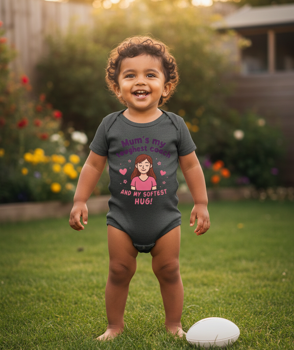 Child wearing a onesie with text and a cartoon character, standing on grass with flowers in the background. The baby is wearing a Cows Corner baby grow with a funny rugby slogan 'Mum’s My Toughest Coach and My Softest Hug!'. Cows Corner gifts are perfect for sport-mad fans, these gifts work brilliantly for birthdays, new baby celebrations, Father’s Day, Mother’s Day, Christmas, anniversaries, thank you gifts, end-of-season team awards, graduations, retirements, and just-because moments