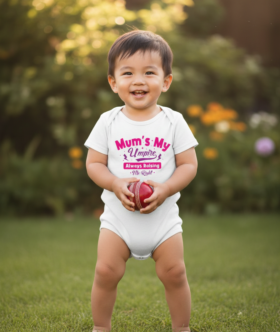Child wearing a white onesie with text, holding a red ball outdoors. The baby is wearing a Cows Corner baby grow with a funny cricket slogan 'Mums my umpire always raising me right'. Cows Corner gifts are perfect for sport-mad fans, these gifts work brilliantly for birthdays, new baby celebrations, Father’s Day, Mother’s Day, Christmas, anniversaries, thank you gifts, end-of-season team awards, graduations, retirements, and just-because moments when you want to raise a smile. 
