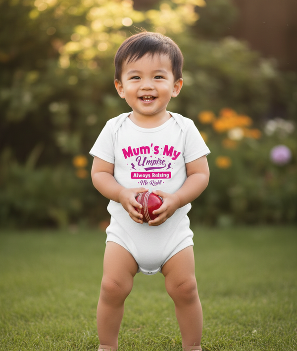 Child wearing a white onesie with text, holding a red ball outdoors. The baby is wearing a Cows Corner baby grow with a funny cricket slogan 'Mums my umpire always raising me right'. Cows Corner gifts are perfect for sport-mad fans, these gifts work brilliantly for birthdays, new baby celebrations, Father’s Day, Mother’s Day, Christmas, anniversaries, thank you gifts, end-of-season team awards, graduations, retirements, and just-because moments when you want to raise a smile. 
