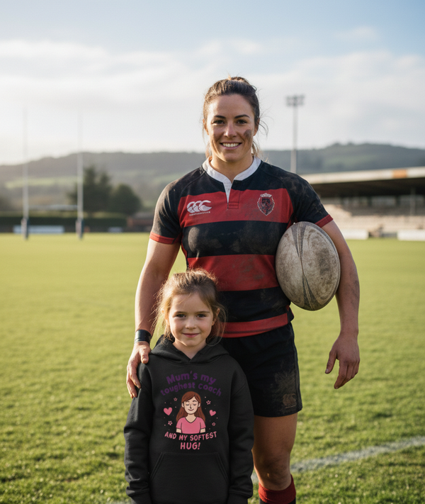 Woman in rugby gear holding a rugby ball with a young girl on a sports field. The girl is wearing a Cows Corner hoodie with the funny rugby slogan 'Mum’s My Toughest Coach and My Softest Hug!'. Cows Corner gifts are perfect for sport-mad fans, these gifts work brilliantly for birthdays, new baby celebrations, Father’s Day, Mother’s Day, Christmas, anniversaries, thank you gifts, end-of-season team awards, graduations, retirements, and just-because moments when you want to raise a smile. 
