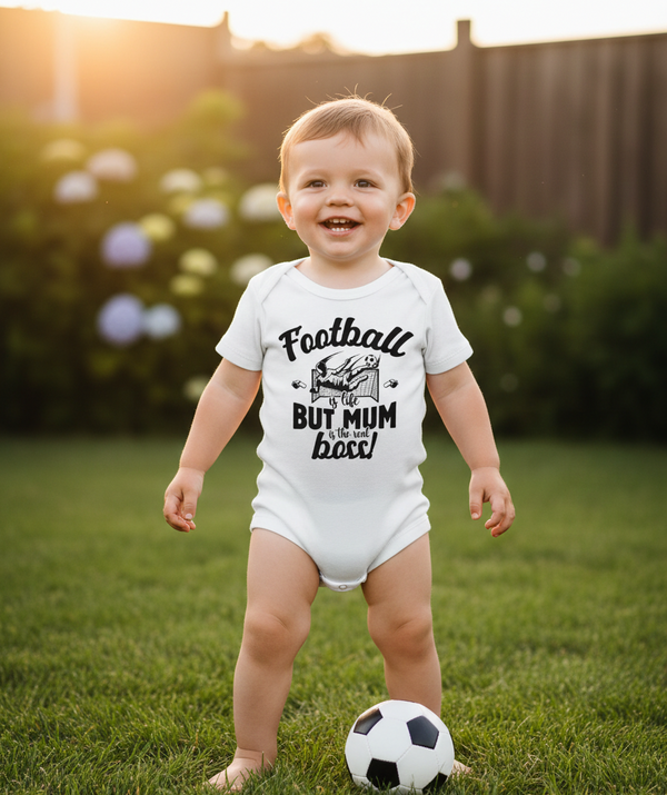 Child wearing a white onesie with text, standing on grass with a soccer ball. The baby is wearing a Cows Corner baby grow with the funny football slogan 'Football Is Life but Mum Is the Real Boss'. Cows Corner gifts are perfect for sport-mad fans, these gifts work brilliantly for birthdays, new baby celebrations, Father’s Day, Mother’s Day, Christmas, anniversaries, thank you gifts, end-of-season team awards, graduations, retirements, and just-because moments when you want to raise a smile. 
