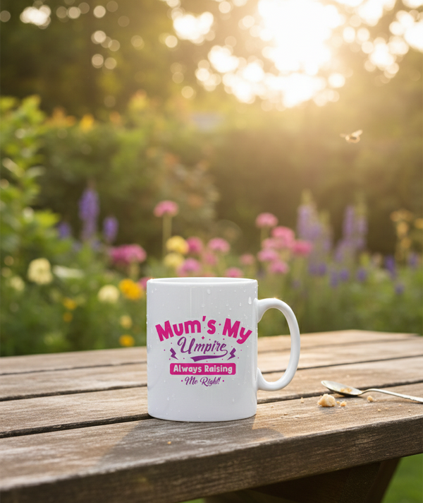A white Cows Corner mug with 'Mum's My Umpire Always Ruling Me Right' text on a wooden table with a garden background. Cows Corner gifts are perfect for sport-mad fans, these gifts work brilliantly for birthdays, new baby celebrations, Father’s Day, Mother’s Day, Christmas, anniversaries, thank you gifts, end-of-season team awards, graduations, retirements, and just-because moments when you want to raise a smile. 
