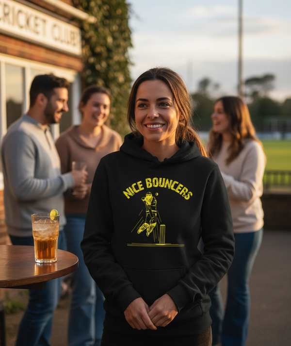 Woman wearing a black Cows Corner hoodie with 'Nice Bouncers' graphic, standing outdoors with people in the background.  Cows Corner gifts are perfect for sport-mad fans, these gifts work brilliantly for birthdays, new baby celebrations, Father’s Day, Mother’s Day, Christmas, anniversaries, thank you gifts, end-of-season team awards, graduations, retirements, and just-because moments when you want to raise a smile. 
