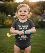 Child wearing a humorous baby onesie with golf-themed text, standing outdoors. The baby is wearing a Cows Corner baby grow with the funny golf slogan 'One Swing, One Hole, One Very Smug Golfer'. Cows Corner gifts are perfect for sport-mad fans, these gifts work brilliantly for birthdays, new baby celebrations, Father’s Day, Mother’s Day, Christmas, anniversaries, thank you gifts, end-of-season team awards, graduations, retirements, and just-because moments when you want to raise a smile. 
