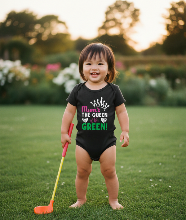 Child wearing a black onesie with text, holding a small golf club in a garden setting.  The baby is wearing a Cows Corner baby grow with a funny golf slogan 'Mum’s the Queen of the Green'. Cows Corner gifts are perfect for sport-mad fans, these gifts work brilliantly for birthdays, new baby celebrations, Father’s Day, Mother’s Day, Christmas, anniversaries, thank you gifts, end-of-season team awards, graduations, retirements, and just-because moments when you want to raise a smile. 
