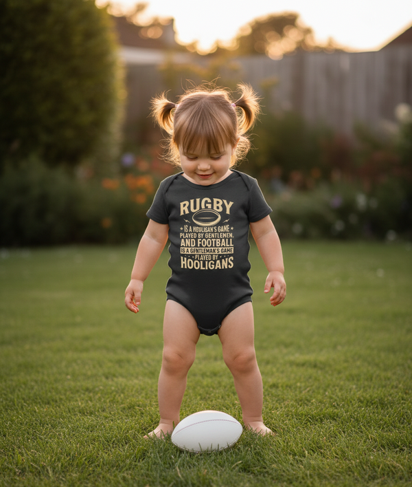 Child wearing a black onesie with text, standing on grass with a rugby ball. The baby is wearing a Cows Corner baby grow with the funny rugby slogan 'Rugby Is a Hooligan’s Game Played by Gentleman, and Football Is a Gentleman’s Game Played by Hooligans'. Cows Corner gifts are perfect for sport-mad fans, these gifts work brilliantly for birthdays, new baby celebrations, Father’s Day, Mother’s Day, Christmas, anniversaries, thank you gifts, end-of-season team awards, graduations, retirements, and just-because