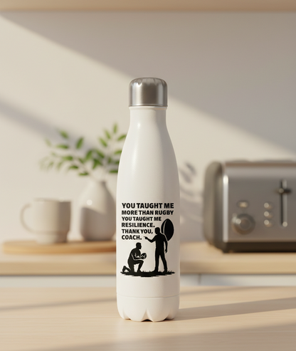 White water bottle with a motivational quote on a kitchen counter. This is a Cows Corner water bottle with a funny rugby slogan 'You Taught Me More Than Rugby, You Taught Me Resilience. Thank You, Coach'. Cows Corner gifts are perfect for sport-mad fans, these gifts work brilliantly for birthdays, new baby celebrations, Father’s Day, Mother’s Day, Christmas, anniversaries, thank you gifts, end-of-season team awards, graduations, retirements, and just-because moments when you want to raise a smile. 
