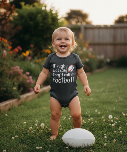 Child wearing a black onesie with a humorous rugby football comparison, standing outdoors in a garden. The baby is wearing a Cows Corner baby grow with a funny rugby slogan 'If Rugby Was Easy They’d Call It Football'. Cows Corner gifts are perfect for sport-mad fans, these gifts work brilliantly for birthdays, new baby celebrations, Father’s Day, Mother’s Day, Christmas, anniversaries, thank you gifts, end-of-season team awards, graduations, retirements, and just-because moments