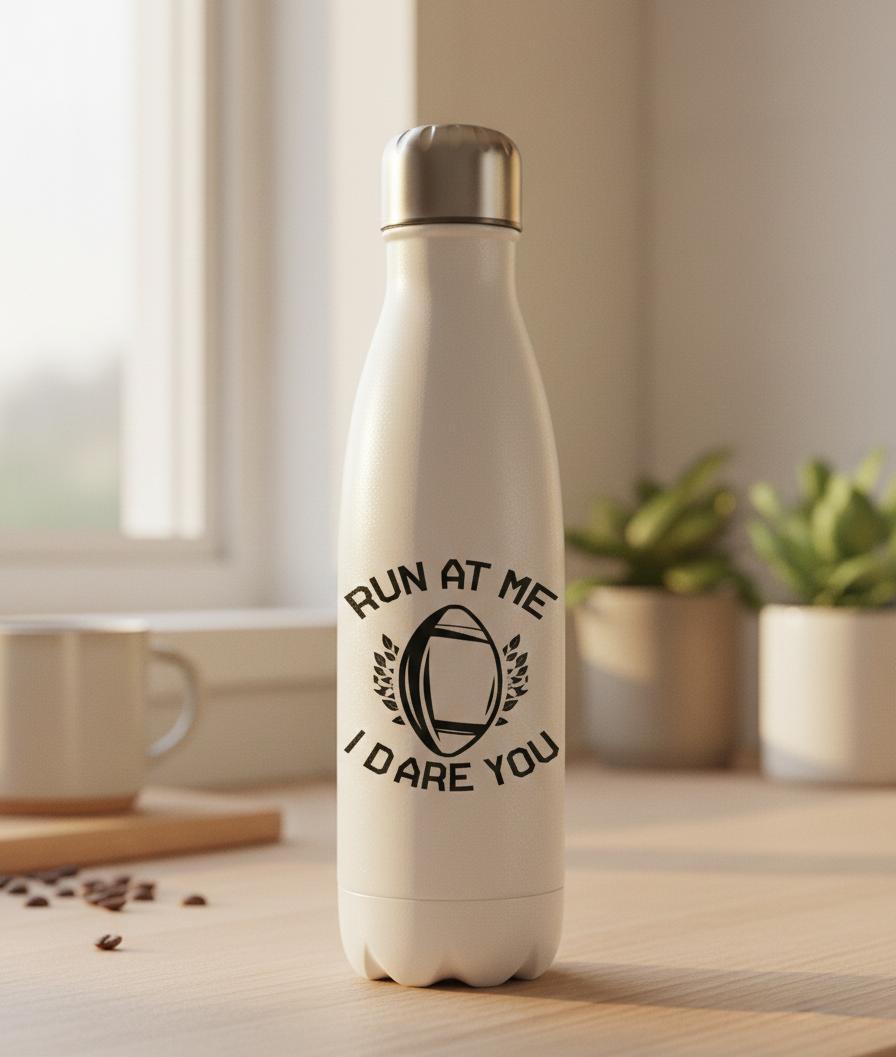 White water bottle with motivational quote on a wooden table with plants in the background. This is a Cows Corner water bottle with a funny rugby slogan 'Run at Me I Dare You'. Cows Corner gifts are perfect for sport-mad fans, these gifts work brilliantly for birthdays, new baby celebrations, Father’s Day, Mother’s Day, Christmas, anniversaries, thank you gifts, end-of-season team awards, graduations, retirements, and just-because moments when you want to raise a smile. 

