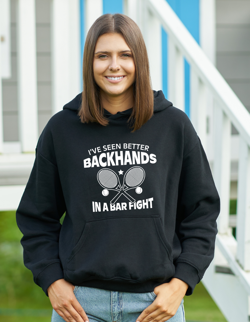 A a lady wearing black hoodie with the funny tennis slogan on the front that reads 'I've Seen Better Backhands In A Bar Fight'