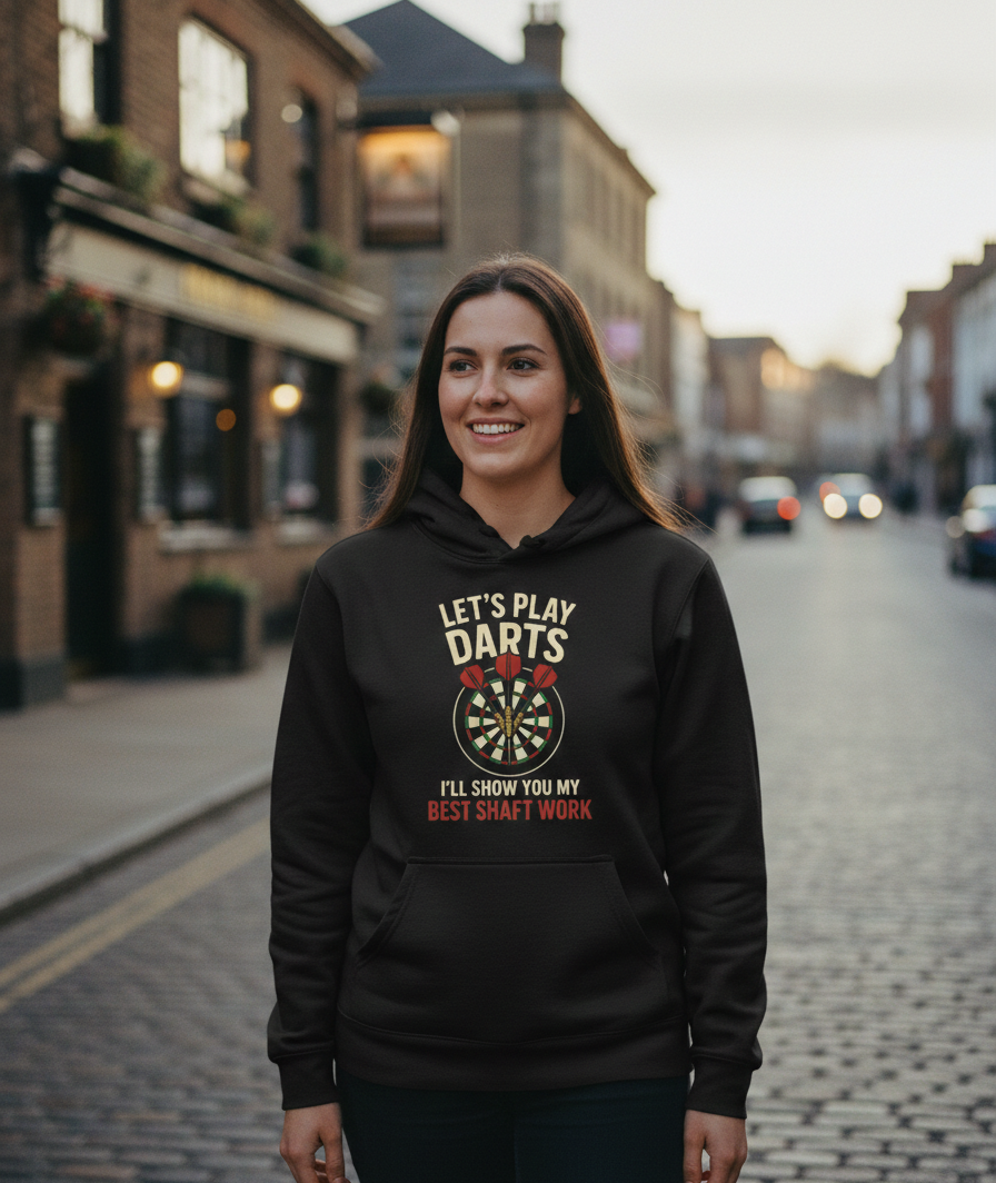 Woman wearing a black hoodie with a dart design and text on a street. The lady is wearing a Cows Corner hoodie with the funny darts slogan 'Lets Play Darts I'll Show You My Best Shaft Work'. Cows Corner gifts are perfect for sport-mad fans, these gifts work brilliantly for birthdays, new baby celebrations, Father’s Day, Mother’s Day, Christmas, anniversaries, thank you gifts, end-of-season team awards, graduations, retirements, and just-because moments when you want to raise a smile. 
