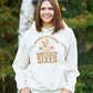 A lady wearing a white hoodie with You Bring the Sledging, I'll Bring the Sixes on the front. A Funny Cricket Hoodie, Premium Fabric, Multiple Adult Sizes