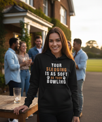 Woman wearing a black hoodie with humorous text, standing outdoors with friends in the background. The lady is wearing a Cows Corner hoodie with the funny cricket slogan 'Your Sledging Is As Soft As Your Bowling'. Cows Corner gifts are perfect for sport-mad fans, these gifts work brilliantly for birthdays, new baby celebrations, Father’s Day, Mother’s Day, Christmas, anniversaries, thank you gifts, end-of-season team awards, graduations, retirements, and just-because moments when you want to raise a smile. 