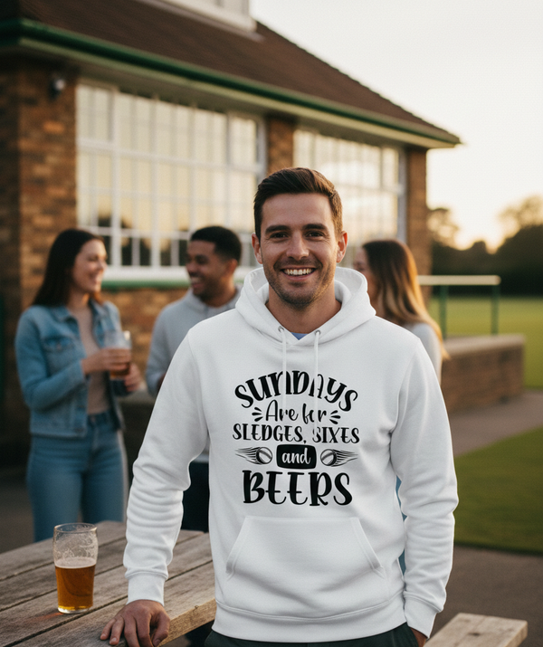 Man wearing a white hoodie with text, standing outdoors with people and a sunset in the background. The man is wearing a Cows Corner hoodie with the funny cricket slogan 'Sundays Are for Sledges, Sixes & Beers'. Cows Corner gifts are perfect for sport-mad fans, these gifts work brilliantly for birthdays, new baby celebrations, Father’s Day, Mother’s Day, Christmas, anniversaries, thank you gifts, end-of-season team awards, graduations, retirements, and just-because moments when you want to raise a smile. 
