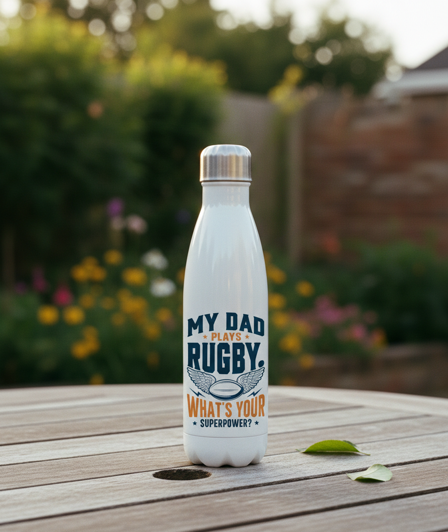 White water bottle with a rugby-themed design on a wooden surface outdoors. This is a Cows Corner water bottle with a funny rugby slogan 'My Dad Plays Rugby. What’s Your Superpower?'. Cows Corner gifts are perfect for sport-mad fans, these gifts work brilliantly for birthdays, new baby celebrations, Father’s Day, Mother’s Day, Christmas, anniversaries, thank you gifts, end-of-season team awards, graduations, retirements, and just-because moments when you want to raise a smile. 

