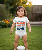 Child wearing a onesie with humorous text outdoors on grass. The baby is wearing a Cows Corner baby grow with a funny rugby slogan 'My Mum Tackles Everything on the Pitch and at Home!'. Cows Corner gifts are perfect for sport-mad fans, these gifts work brilliantly for birthdays, new baby celebrations, Father’s Day, Mother’s Day, Christmas, anniversaries, thank you gifts, end-of-season team awards, graduations, retirements, and just-because moments when you want to raise a smile. 
