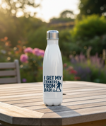 White water bottle with text and graphic on a wooden table outdoors. This is a Cows Corner water bottle with a funny football slogan 'I Get My Tekkers From Dad'. Cows Corner gifts are perfect for sport-mad fans, these gifts work brilliantly for birthdays, new baby celebrations, Father’s Day, Mother’s Day, Christmas, anniversaries, thank you gifts, end-of-season team awards, graduations, retirements, and just-because moments when you want to raise a smile. 
