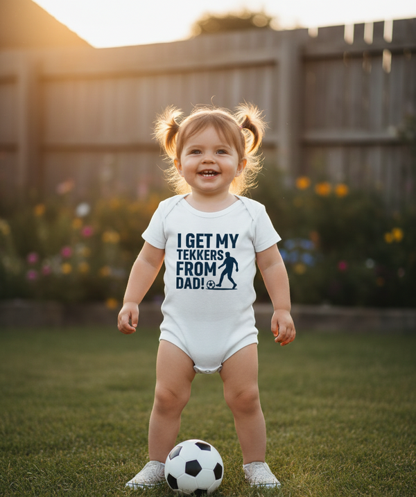 Child wearing a onesie with text standing on grass with a soccer ball. The baby is wearing a Cows Corner baby grow with a funny football slogan 'I Get My Tekkers From Dad'. Cows Corner gifts are perfect for sport-mad fans, these gifts work brilliantly for birthdays, new baby celebrations, Father’s Day, Mother’s Day, Christmas, anniversaries, thank you gifts, end-of-season team awards, graduations, retirements, and just-because moments when you want to raise a smile. 

