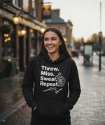 Person wearing a black hoodie with text and graphics on a street. The lady is wearing a Cows Corner hoodie with the funny darts slogan 'Throw. Miss. Swear. Repeat'. Cows Corner gifts are perfect for sport-mad fans, these gifts work brilliantly for birthdays, new baby celebrations, Father’s Day, Mother’s Day, Christmas, anniversaries, thank you gifts, end-of-season team awards, graduations, retirements, and just-because moments when you want to raise a smile. 

