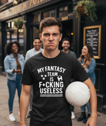 Man holding a soccer ball wearing a t-shirt with a humorous message, standing in front of a pub with people in the background. A Cows Corner t-shirt with the funny football slogan 'My Fantasy Team Is F*cking Useless'. Cows Corner gifts are perfect for sport-mad fans, these gifts work brilliantly for birthdays, new baby celebrations, Father’s Day, Mother’s Day, Christmas, anniversaries, thank you gifts, end-of-season team awards, graduations, retirements, and just-because moments when you want to smile