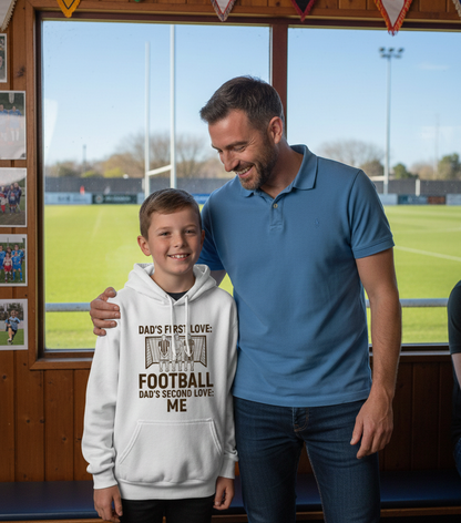 Man and young boy standing together in a sports facility, with the boy wearing a hoodie with text. The boy is wearing a Cows Corner hoodie with the funny football slogan 'Dad’s First Love: Football. Dad’s Second Love: Me'. Cows Corner gifts are perfect for sport-mad fans, these gifts work brilliantly for birthdays, new baby celebrations, Father’s Day, Mother’s Day, Christmas, anniversaries, thank you gifts, end-of-season team awards, graduations, retirements, and just-because moments 